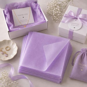Può includere: Una pila di fogli di carta velina color viola chiaro, una scatola regalo bianca aperta con una collana d'oro su un biglietto di ringraziamento, una scatola chiusa con nastro viola, un sacchetto viola con coulisse e un piattino in ceramica.