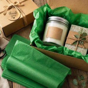 Può includere: Una scatola regalo contenente una candela bianca in un barattolo di vetro con etichetta marrone vuota e una saponetta rettangolare maculata avvolta in carta marrone con un fiocco di spago e una piccola foglia, adagiata su carta velina verde.
