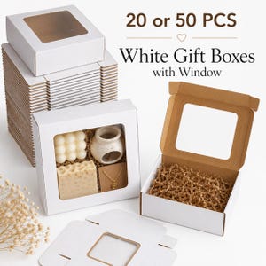 Op de afbeelding: Een stapel witte geschenkdozen met venster, voorzien van de tekst 20 or 50 PCS White Gift Boxes with Window. Eén doos is geopend en gevuld met bruin opvulmateriaal, een andere is gesloten en toont een kaars, zeep en een gouden ketting.
