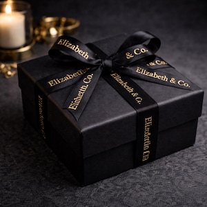 May include: A black gift box tied with a black ribbon featuring the text "Elizabeth & Co." in gold. The box is square and sits on a dark surface. A lit candle and gold decorative items are in the background.