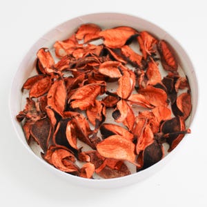 May include: A white, circular bowl filled with dried, orange-red potpourri. The potpourri pieces have a textured, leaf-like appearance, with some showing darker, brown edges. The arrangement is set against a plain white background.