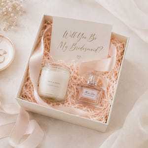 Peut inclure: Une boîte cadeau blanche remplie de papier déchiqueté rose clair, contenant une bougie en pot en verre étiquetée Rose & Peony, un flacon de parfum carré en verre étiqueté Miss Dior Blooming Bouquet et une carte avec le texte Will You Be My Bridesmaid?