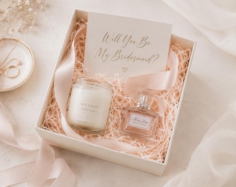 May include: A white gift box filled with light pink paper shred, containing a glass jar candle labeled Rose & Peony, a square glass perfume bottle labeled Miss Dior Blooming Bouquet, and a card with the text Will You Be My Bridesmaid?