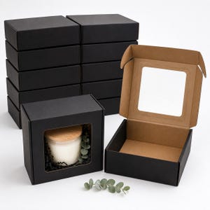 May include: A collection of rectangular black cardboard boxes with clear plastic windows. One box is open, revealing a natural brown interior. Another box contains a white candle with a wooden lid and green foliage, surrounded by black filler.