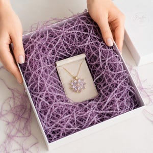 Pode incluir: Um colar dourado com um pendente em forma de flor com pedras roxas claras em corte marquise, apresentado numa caixa quadrada branca cheia de papel picado roxo. O pendente repousa sobre um pequeno cartão de exposição bege na caixa.