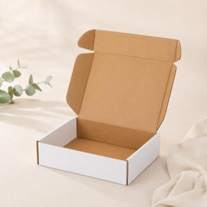 May include: An open, square cardboard box with a white exterior and a brown interior. The box has flaps that are partially open. The background is a light beige color with a sprig of green leaves.