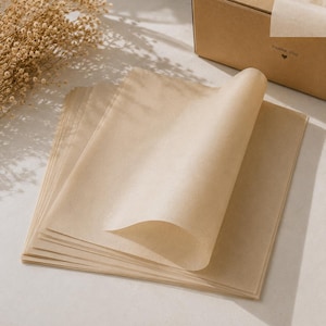 Pode incluir: Uma pilha de folhas de papel de seda retangulares e translúcidas de cor castanho-claro. A folha superior está parcialmente enrolada para cima. Ao lado do papel, encontra-se uma caixa de cartão castanha com o texto THANK YOU e um coração.