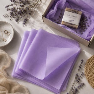 Può includere: Una pila di fogli di carta velina color viola chiaro. Nelle vicinanze si trova una scatola regalo in cartone aperta contenente una saponetta con l'etichetta Lavender, un rametto di lavanda essiccata, un piattino in ceramica con anelli dorati e un tessuto beige.