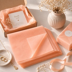 Può includere: Una pila di fogli di carta velina color pesca accanto a una scatola di cartone aperta contenente un biglietto di ringraziamento con un'icona a forma di cuore, un vaso in ceramica a coste con fiori secchi, un regalo incartato e un nastro.