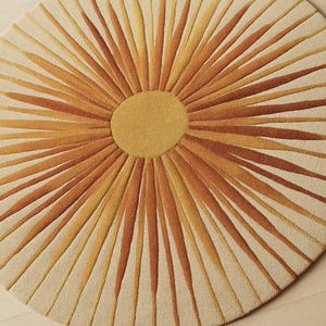 Puede incluir: Una alfombra redonda con dise&ntilde;o de sol. Presenta un c&iacute;rculo central amarillo con rayos naranjas y amarillos sobre un fondo crema. La alfombra est&aacute; hecha de un material suave y texturizado. Di&aacute;metro aproximado de 150 cm.
