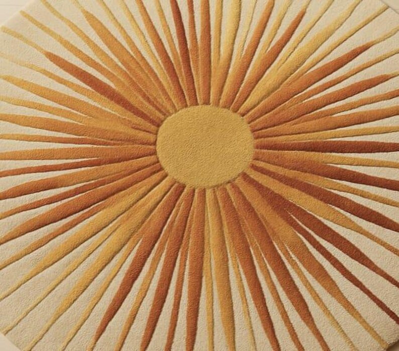 Puede incluir: Una alfombra redonda con un dise&ntilde;o de sol. El centro es de color amarillo-dorado s&oacute;lido, con l&iacute;neas radiantes en tonos naranja y amarillo-anaranjado sobre un fondo crema. La alfombra tiene un aspecto suave y texturizado.