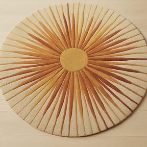 Puede incluir: Una alfombra redonda con dise&ntilde;o de rayos de sol. La alfombra presenta un c&iacute;rculo central amarillo mostaza, con l&iacute;neas radiantes en tonos naranja y beige sobre un fondo crema. Un accesorio decorativo para el hogar, de unos 150 cm de di&aacute;metro.
