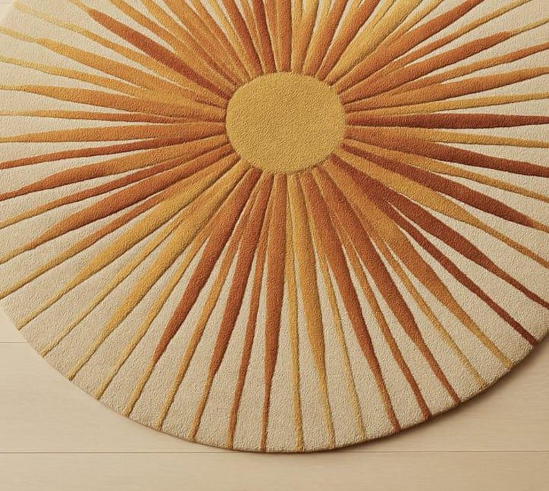 Puede incluir: Una alfombra redonda con dise&ntilde;o de sol. La alfombra presenta un c&iacute;rculo central amarillo con l&iacute;neas radiantes en tonos naranja y marr&oacute;n sobre un fondo crema. La alfombra est&aacute; hecha de un material suave y texturizado.
