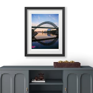 May include: A framed photograph of a bridge over a river with a reflection of the bridge in the water. The sky is blue with white clouds. The bridge is green and has a curved arch. The water is calm and reflects the sky and the bridge.