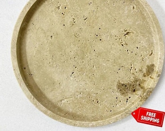 May include: A round, beige travertine stone tray with a raised rim. The tray has a natural, rustic look with a mottled surface.