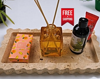 May include: A rectangular, light brown, travertine tray with a wavy edge. The tray contains a small, square, amber glass bottle with wooden sticks, a small, rectangular, pink and orange box with a peach design, a small, clear glass bottle with a black lid, and a small, red and white tube with a pink flower design. The text on the pink and orange box reads "Lemon Home". The text on the clear glass bottle reads "Organic Olive". The text on the red and white tube reads "farm kid".