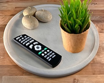 May include: A white round tray with a black remote control, three beige decorative stones, and a potted green plant with brown paper wrapping around the pot.