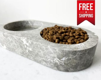 May include: A double pet bowl with a gray and white marble pattern. One side of the oval-shaped bowl is empty, while the other side is filled with brown, round dry pet food kibble. The surface has a smooth, polished stone appearance.