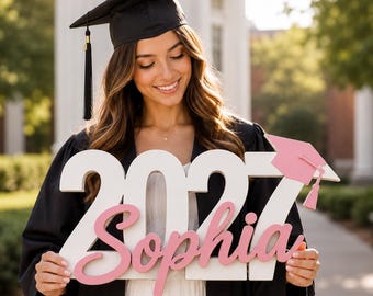 May include: A white wooden cutout of the number 2027 with the name Sophia in pink script lettering attached to the front. A small pink graduation cap icon is attached to the top right of the number seven. The sign is held by a graduate.