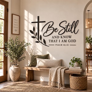 May include: A black metal wall art piece featuring a cross with a leafy branch and the text Be Still AND KNOW THAT I AM GOD PSALM 46:10. The sign is mounted on a light-colored wall above a wooden bench with pillows and a throw blanket.