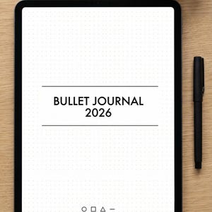May include: A digital tablet showing a bullet journal layout for 2026, with a dotted grid and the text "BULLET JOURNAL 2026". A black pen lies next to the tablet on a wooden surface, with a white mug of coffee in the background.