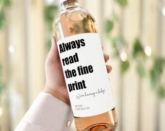 May include: A clear wine bottle with a pink label that reads "Always read the fine print. We're having a baby!" The bottle has a pink cap and is filled with a light pink liquid. The text "Baby due: Coming August 2024" is also on the label.