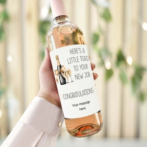 Puede incluir: Una botella de vino blanco con una etiqueta que dice "Aquí tienes un pequeño brindis por tu nuevo trabajo. ¡Felicidades! Tu mensaje aquí." La etiqueta también presenta una cinta dorada y dos copas de champán.