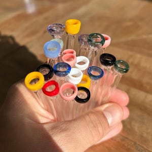 Puede incluir: Un mont&oacute;n de pipas para fumar de vidrio con anillos de vidrio de colores en el extremo. Las pipas son de vidrio transparente y los anillos son de varios colores, incluyendo amarillo, rojo, azul, verde y blanco.