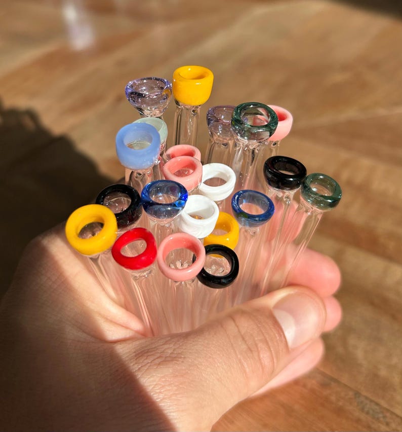 Puede incluir: Un mont&oacute;n de pipas para fumar de vidrio con anillos de vidrio de colores en el extremo. Las pipas son de vidrio transparente y los anillos son de varios colores, incluyendo amarillo, rojo, azul, verde y blanco.