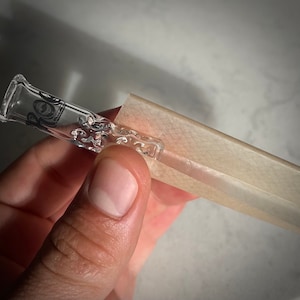 Può includere: Un filtro in vetro trasparente con vortici e motivi decorativi è tenuto accanto a una carta beige. Il filtro ha un'estremità svasata e un lungo stelo sottile. Il design è visibile attraverso il vetro trasparente.