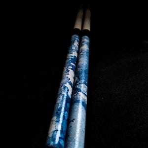 May include: A pair of wooden drumsticks with a blue and white patterned design. The drumsticks have black rubber tips.