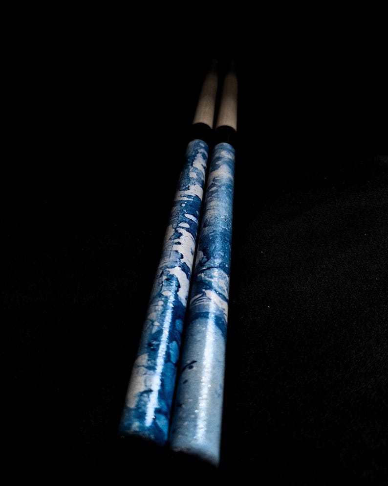 May include: A pair of wooden drumsticks with a blue and white patterned design. The drumsticks have black rubber tips.