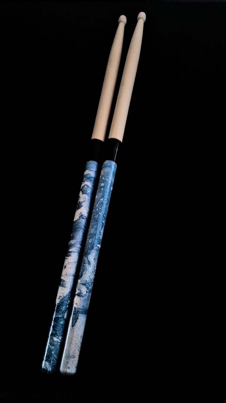 May include: Two wooden drumsticks with a blue and white marbled design. The drumsticks have black tips.