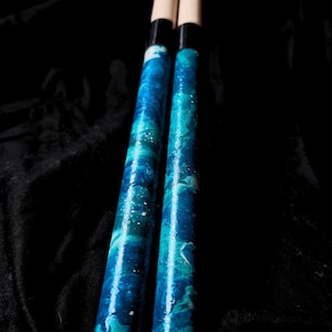 May include: Two drumsticks with blue and white marbled shafts and natural wood tips. The shafts have a glossy finish with a swirling pattern of blues and white. The tips are a light beige color.