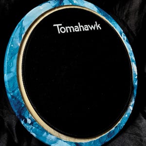 Op de afbeelding: Ronde drum oefenpad met een zwart speeloppervlak en het woord "Tomahawk" in het wit. De rand heeft een blauw-wit marmerpatroon, met een beige gekleurde rand.