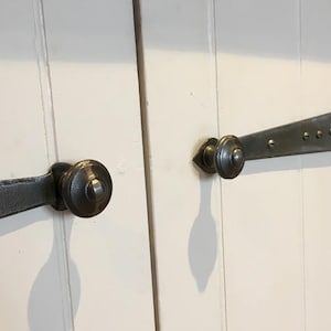 May include: Close-up of white wooden doors featuring antique metal hardware. The hardware includes a hinge and a latch with a round knob. The metal has a dark, aged finish, and the doors are painted white.