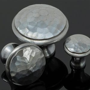 May include: A selection of silver cabinet knobs with a textured, hammered finish. The knobs are in various sizes, with a large, rounded knob at the center and smaller ones around it. The metal has a reflective surface.