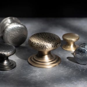 May include: A selection of cabinet knobs in metallic finishes. The knobs have hammered and smooth textures in silver, bronze, and gold. The knobs are displayed on a grey surface.