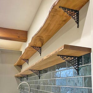 May include: Two wooden shelves with live edges are mounted on a wall. The shelves are supported by black, decorative metal brackets. The wall is painted a light colour and has a section of green tile.