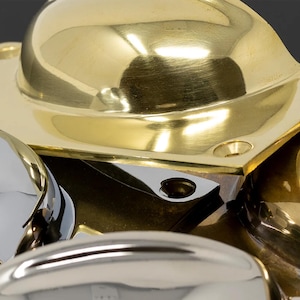 May include: Close-up of various shiny metal hardware components. The image shows a polished brass cup-shaped element with a square base, alongside other rounded, silver-coloured pieces. The hardware has a reflective surface.