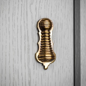 May include: Close-up of a brass keyhole cover with an ornate, elongated design. The cover is mounted on a light grey wooden door. The keyhole cover features horizontal ridges and a rounded top.