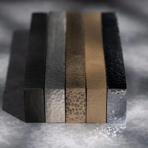 May include: Five rectangular metal bars with textured surfaces are displayed. The bars are in shades of black, silver, bronze, and gold. The lighting creates shadows and highlights the different textures and colours.