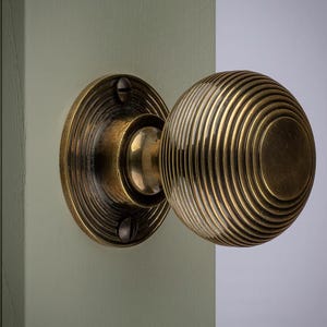 May include: Close-up of a brass door knob with a round, grooved design. The knob is mounted on a light green door. The circular backplate is also brass and has a ridged pattern. The hardware has a vintage aesthetic.