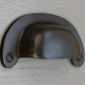May include: A dark grey, cup-style drawer pull with a semi-circular design. The metal hardware features a curved handle and is attached with two screws. The finish has a brushed appearance, suggesting a vintage or industrial aesthetic.