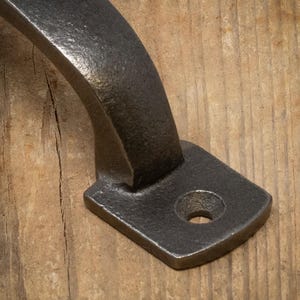 May include: Close-up of a dark grey, cast iron handle. The handle has a curved design with a rectangular base and a single hole for mounting. The surface has a textured, rustic appearance, suggesting a vintage or industrial style.