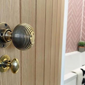 K&ouml;nnte beinhalten: Nahaufnahme eines T&uuml;rknaufs aus Messing an einer hellen Holzt&uuml;r. Der Hauptknauf hat ein rundes, geripptes Design, und der kleinere Knauf darunter ist oval. Der Hintergrund zeigt eine rosa geflieste Wand und eine wei&szlig;e Badewanne.
