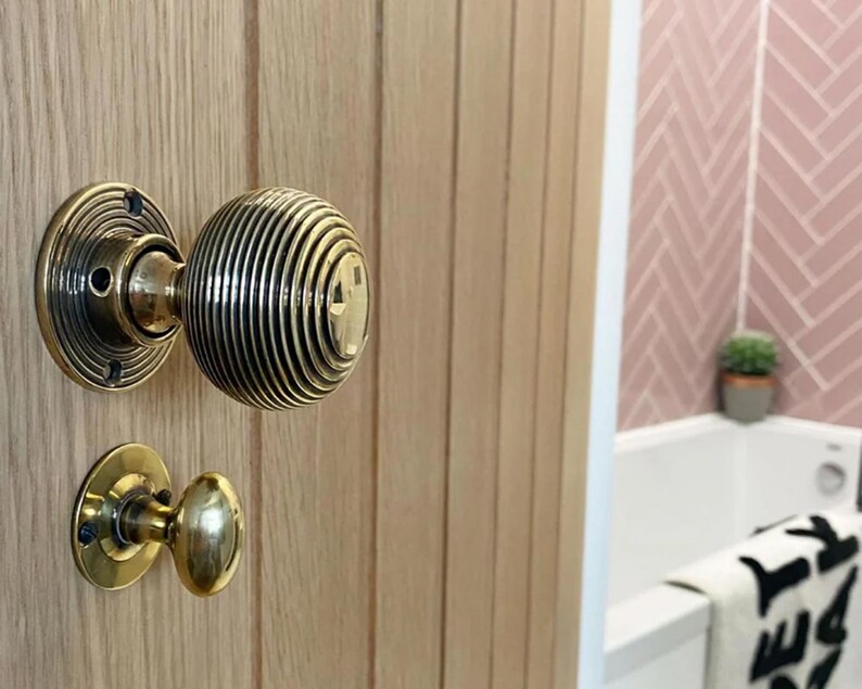 K&ouml;nnte beinhalten: Nahaufnahme eines T&uuml;rknaufs aus Messing an einer hellen Holzt&uuml;r. Der Hauptknauf hat ein rundes, geripptes Design, und der kleinere Knauf darunter ist oval. Der Hintergrund zeigt eine rosa geflieste Wand und eine wei&szlig;e Badewanne.