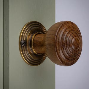 May include: A wooden door knob with a brass backplate. The knob is a warm brown colour with concentric circular grain patterns. The backplate is a shiny gold colour. The knob is attached to a light green door.