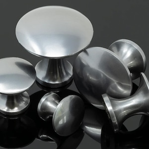 May include: A selection of brushed nickel cabinet knobs in different sizes. The knobs feature a rounded, mushroom-shaped design with a smooth, metallic finish. The knobs are arranged on a reflective surface, creating light and shadow effects.
