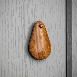 May include: A teardrop-shaped wooden cabinet knob with a brass screw. The knob is made of a warm-toned wood and is mounted on a grey cabinet door. The wood grain is visible.